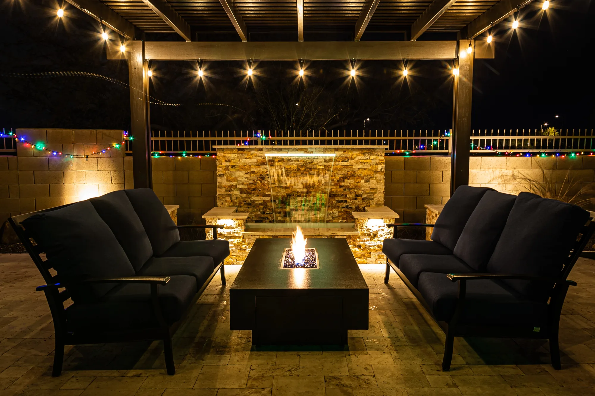 Pergola-covered firepit lounge with a stone water feature backdrop at night