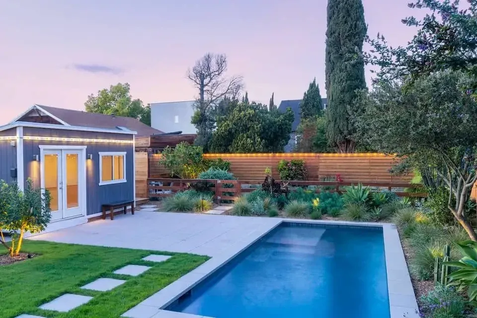 Backyard pool at dusk with timber fencing, garden bed planting and a garden studio